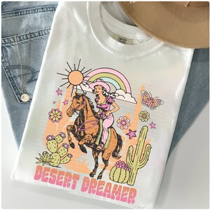 Pu&ograve; includere: Maglietta bianca con una stampa grafica di una donna a cavallo in un paesaggio desertico. La grafica include un arcobaleno, un cactus, stelle e il testo "DESERT DREAMER".