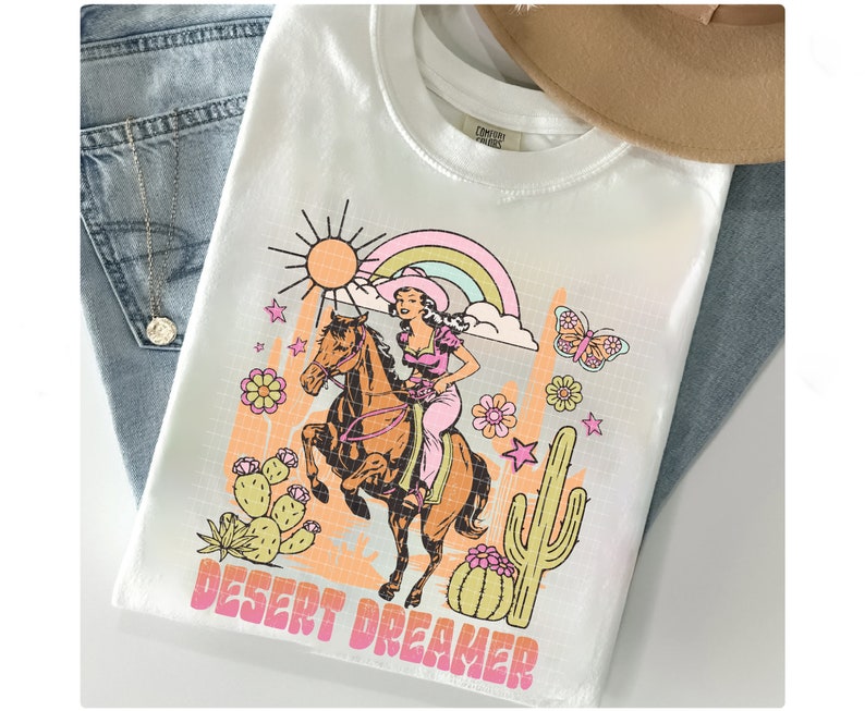 Pu&ograve; includere: Maglietta bianca con una stampa grafica di una donna a cavallo in un paesaggio desertico. La grafica include un arcobaleno, un cactus, stelle e il testo "DESERT DREAMER".