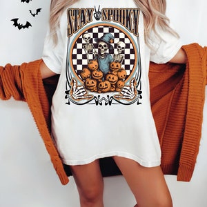 Puede incluir: Camiseta blanca con un gr&aacute;fico de un esqueleto con un sombrero de bruja y sosteniendo una calabaza. El gr&aacute;fico est&aacute; rodeado de un c&iacute;rculo a cuadros y el texto "Stay Spooky".