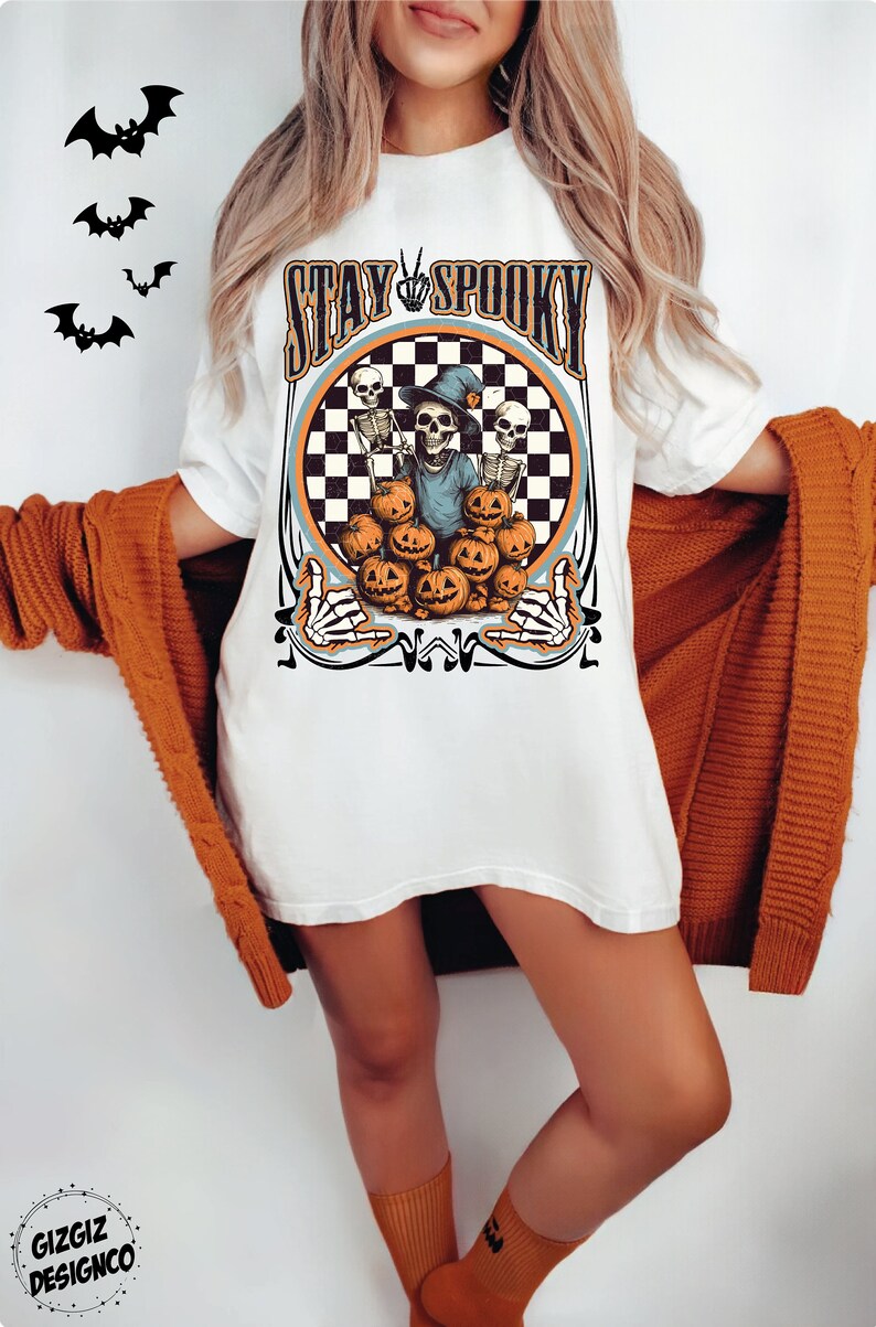 Puede incluir: Camiseta blanca con un gr&aacute;fico de un esqueleto con un sombrero de bruja y sosteniendo una calabaza. El gr&aacute;fico est&aacute; rodeado de un c&iacute;rculo a cuadros y el texto "Stay Spooky".