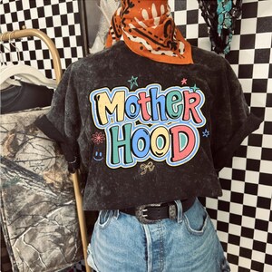 May include: A dark grey, cropped t-shirt with the word "Motherhood" in colourful, bubble letters. The shirt has rolled-up sleeves and is paired with denim shorts and a black belt. An orange bandana is worn on top.