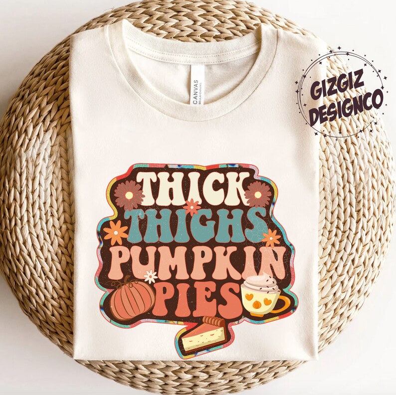 May include: A white t-shirt featuring a retro-style graphic that reads "Thick Thighs Pumpkin Pies". The graphic includes a pumpkin, a cup of coffee, and a slice of pie.