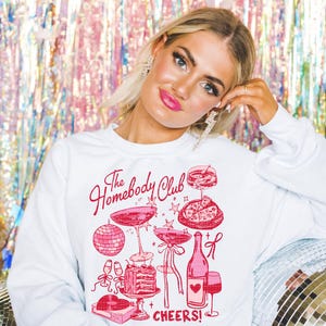 May include: White sweatshirt with a pink graphic design that says "The Homebody Club" and features a disco ball, wine glasses, a bottle of wine, a slice of pizza, a cake, and the text "Cheers!"