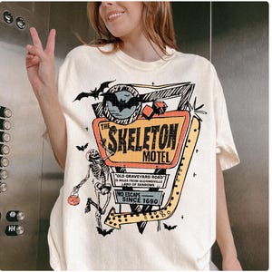 Puede incluir: Camiseta color crema con un gr&aacute;fico colorido del letrero "The Skeleton Motel", un esqueleto, murci&eacute;lagos y el texto "Old Graveyard Road" y "No Escape Since 1690". El dise&ntilde;o tiene acentos naranjas, negros y amarillos.