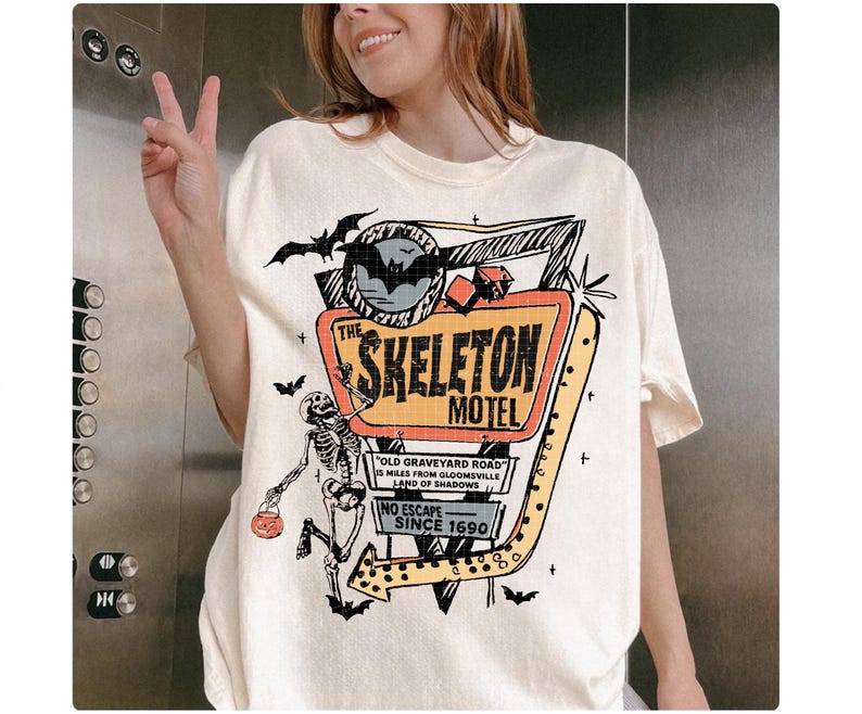 Puede incluir: Camiseta color crema con un gr&aacute;fico colorido del letrero "The Skeleton Motel", un esqueleto, murci&eacute;lagos y el texto "Old Graveyard Road" y "No Escape Since 1690". El dise&ntilde;o tiene acentos naranjas, negros y amarillos.