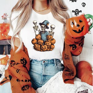 Puede incluir: Una camiseta blanca con un gr&aacute;fico de tres esqueletos con sombreros de bruja y sosteniendo calabazas. Los esqueletos est&aacute;n rodeados de calabazas.