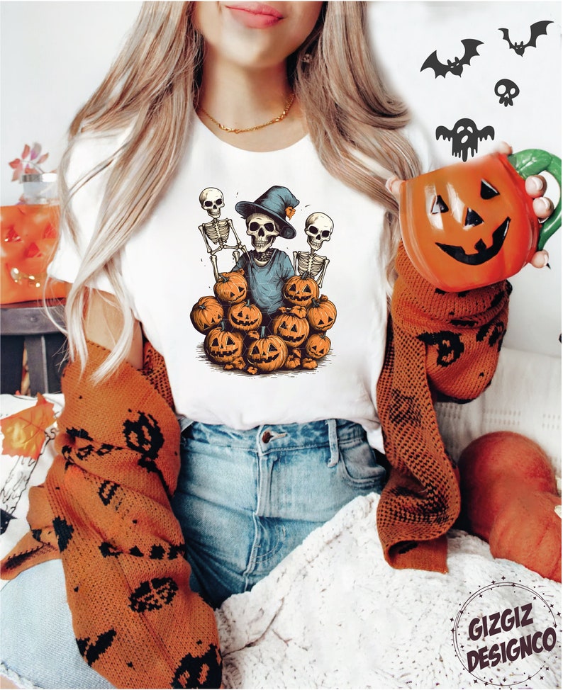 Puede incluir: Una camiseta blanca con un gr&aacute;fico de tres esqueletos con sombreros de bruja y sosteniendo calabazas. Los esqueletos est&aacute;n rodeados de calabazas.