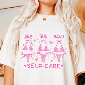 May include: White t-shirt with a pink graphic of three bikini tops and bottoms, with the words "SEA SUN SAND" above and "SELF-CARE" below. The bikini designs vary, with one featuring a zebra print.