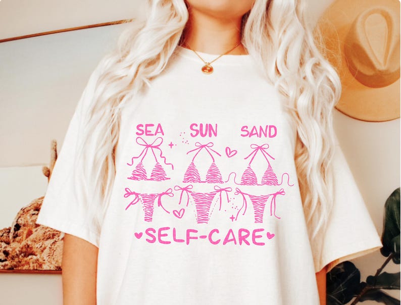 May include: White t-shirt with a pink graphic of three bikini tops and bottoms, with the words "SEA SUN SAND" above and "SELF-CARE" below. The bikini designs vary, with one featuring a zebra print.