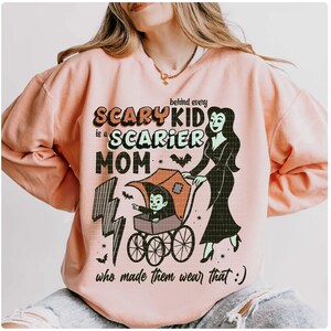 May include: A light peach-colored sweatshirt with a Halloween-themed graphic. The graphic features the text "Behind every scary kid is a scarier mom" with a cartoon image of a vampire child in a stroller being pushed by a vampire woman.