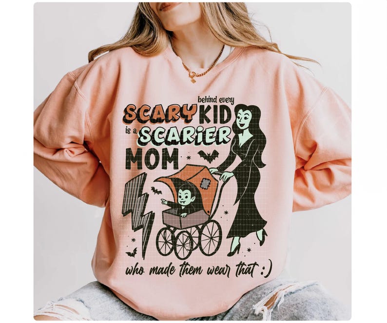 May include: A light peach-colored sweatshirt with a Halloween-themed graphic. The graphic features the text "Behind every scary kid is a scarier mom" with a cartoon image of a vampire child in a stroller being pushed by a vampire woman.