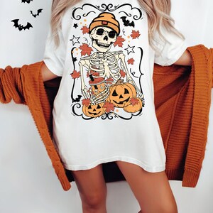 May include: White t-shirt with a graphic of a skeleton wearing a beanie and holding pumpkins. The skeleton is surrounded by fall leaves and stars.