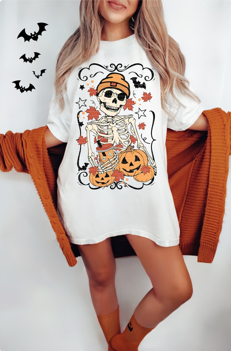 May include: White t-shirt with a graphic of a skeleton wearing a beanie and holding pumpkins. The skeleton is surrounded by fall leaves and stars.