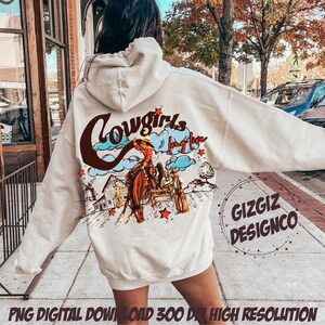 May include: Cream-colored hoodie with a western-themed graphic. The design features a cowgirl on a horse, a windmill, and the words "Cowgirls long live" in a brown and blue color scheme. The hoodie also has the text "GIZGIZ DESIGNCO".