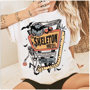 Puede incluir: Camiseta blanca con un dise&ntilde;o retro de "The Skeleton Motel". El letrero es naranja, amarillo y negro, con un esqueleto y murci&eacute;lagos. El texto incluye "Old Graveyard Road" y "Since 1690."