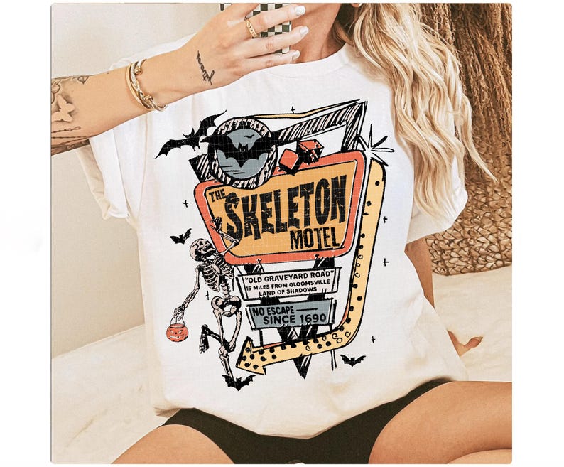 Puede incluir: Camiseta blanca con un dise&ntilde;o retro de "The Skeleton Motel". El letrero es naranja, amarillo y negro, con un esqueleto y murci&eacute;lagos. El texto incluye "Old Graveyard Road" y "Since 1690."