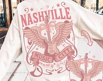 Op de afbeelding: Crèmekleurige sweatshirt met een roze grafisch ontwerp. Het ontwerp toont het woord "Nashville" boven een gevleugelde gitaar, met de woorden "Music City" op een banner. Muzieknoten en sterren omringen de gitaar.