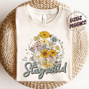 May include: A cream-colored t-shirt with a floral design and the text "Stay Wild" in blue lettering. The floral design features a variety of flowers in shades of yellow, pink, and white, arranged in a circle.