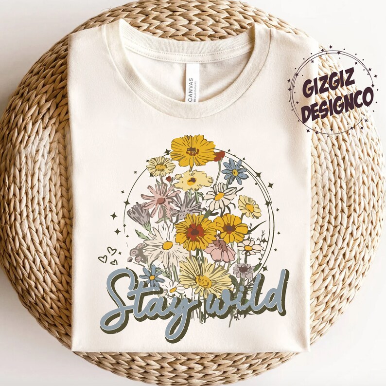 May include: A cream-colored t-shirt with a floral design and the text "Stay Wild" in blue lettering. The floral design features a variety of flowers in shades of yellow, pink, and white, arranged in a circle.