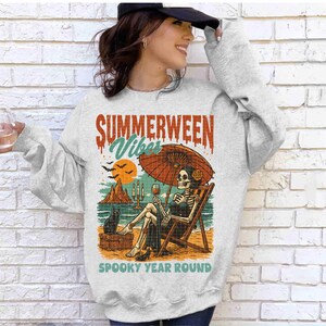 May include: A light gray sweatshirt with a graphic that reads "Summerween Vibes Spooky Year Round." The graphic depicts a skeleton relaxing on a beach chair under an umbrella, with a black cat and a glass of wine.