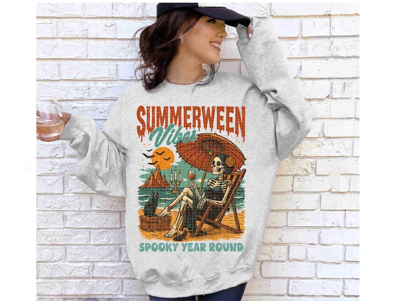May include: A light gray sweatshirt with a graphic that reads "Summerween Vibes Spooky Year Round." The graphic depicts a skeleton relaxing on a beach chair under an umbrella, with a black cat and a glass of wine.