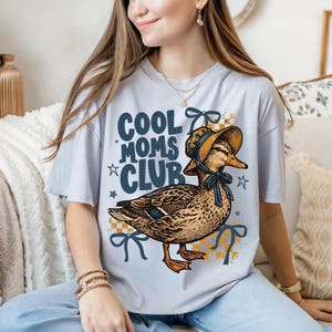 May include: A light blue t-shirt with the text "COOL MOMS CLUB" and a graphic of a duck wearing a bonnet. The duck is brown and tan with orange feet. The shirt has blue bows and stars.