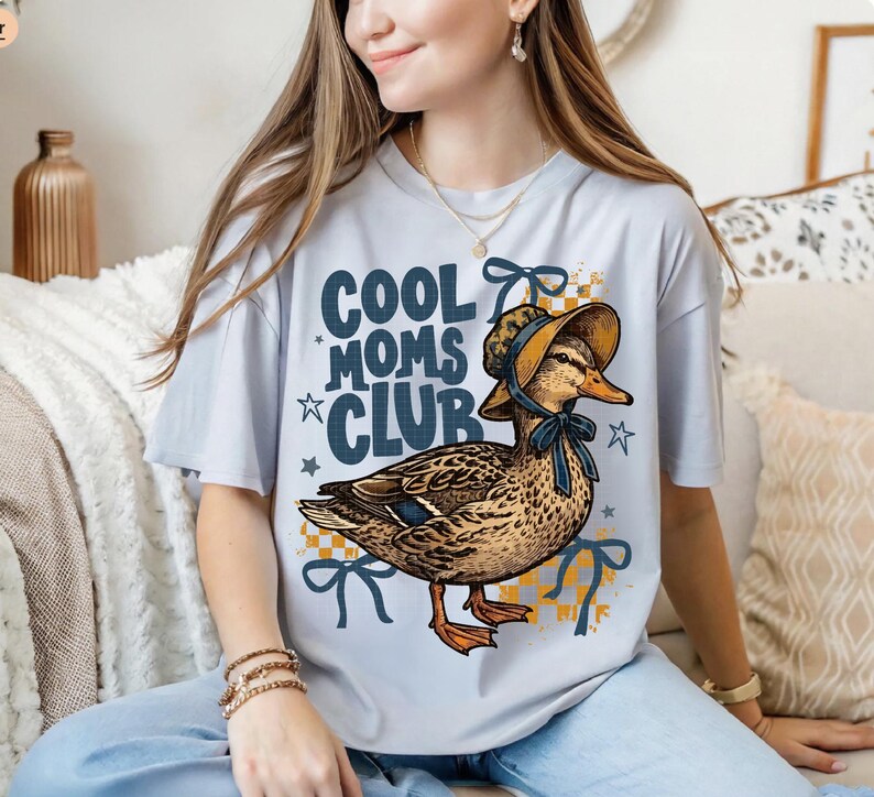 May include: A light blue t-shirt with the text "COOL MOMS CLUB" and a graphic of a duck wearing a bonnet. The duck is brown and tan with orange feet. The shirt has blue bows and stars.