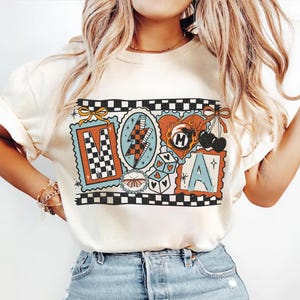Op de afbeelding: Cr&egrave;mekleurig T-shirt met een retro grafisch ontwerp. Het ontwerp bevat de letters "M" en "A" in postzegelstijl frames, een bliksemschicht, dobbelstenen, een hart en kersen. Het shirt heeft een geblokte rand.