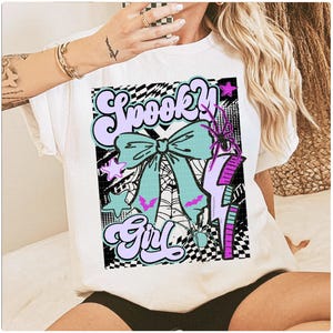 May include: White t-shirt featuring a colorful graphic design. The design includes the words "Spooky Girl" in a retro font, a teal bow, a spider web, a purple lightning bolt, and a spider. The design has a checkerboard background.