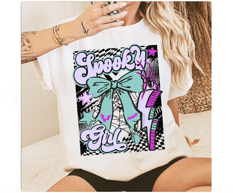 May include: White t-shirt featuring a colorful graphic design. The design includes the words "Spooky Girl" in a retro font, a teal bow, a spider web, a purple lightning bolt, and a spider. The design has a checkerboard background.
