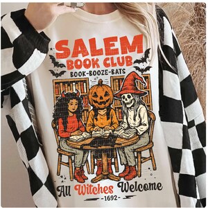 Może przedstawiać: Kremowa bluza z nadrukiem z napisem "Salem Book Club" oraz ilustracjami czarownic i nietoperzy. Projekt zawiera postać z dyniową głową, szkielet i postać z rogami czytające książki wok&oacute;ł stołu. Obecne jest r&oacute;wnież zdanie "All Witches Welcome - 1692".