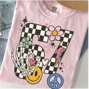 May include: A light pink t-shirt with a retro-style graphic featuring the number 67 in a checkered pattern. The design includes a skeleton hand, smiley face, flowers, hearts, a peace sign, and a lightning bolt.