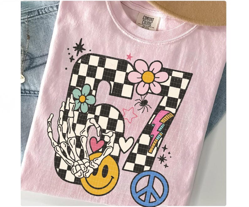 May include: A light pink t-shirt with a retro-style graphic featuring the number 67 in a checkered pattern. The design includes a skeleton hand, smiley face, flowers, hearts, a peace sign, and a lightning bolt.