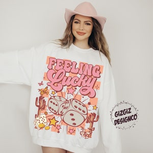 May include: White sweatshirt with the text "Feeling Lucky" in pink, featuring dice, stars, and cacti. The design includes butterflies and playing cards. The sweatshirt has a crew neck and long sleeves.