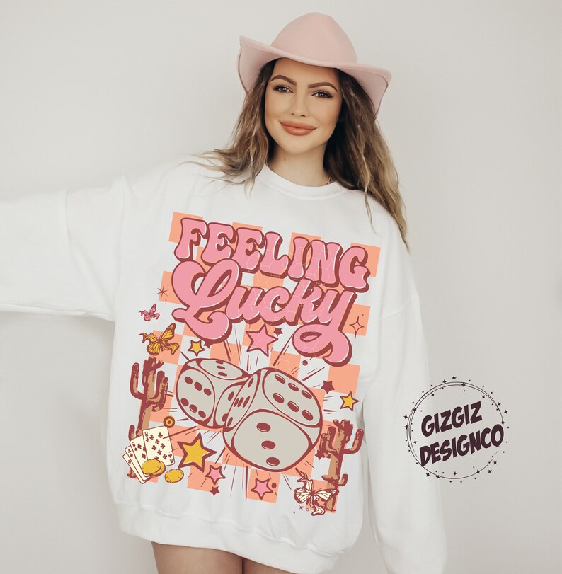 May include: White sweatshirt with the text "Feeling Lucky" in pink, featuring dice, stars, and cacti. The design includes butterflies and playing cards. The sweatshirt has a crew neck and long sleeves.