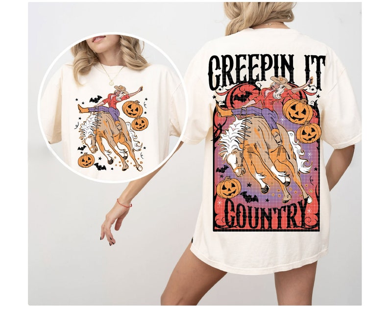 Puede incluir: Una camiseta blanca con un estampado gr&aacute;fico de una mujer montando un caballo con calabazas y murci&eacute;lagos. El texto "Creepin It Country" est&aacute; impreso en la camiseta.