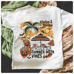 May include: White t-shirt with a colorful graphic featuring a beach scene with a palm tree, sun, and text that reads "Chillin' & Thrillin'" and "Summer-ween Vibes". The design includes jack-o'-lantern strawberries, a beach umbrella, and a slice of watermelon.