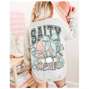 Puede incluir: Una sudadera gris claro con un estampado gr&aacute;fico de conchas marinas y un lazo. El texto "Salty Soul" est&aacute; impreso en la sudadera.
