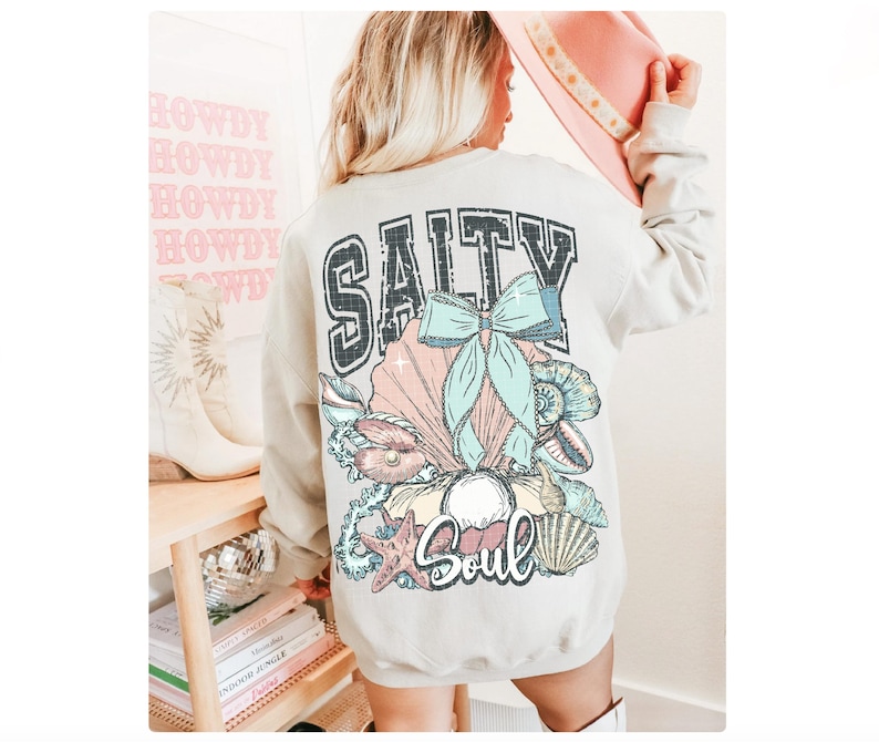 Puede incluir: Una sudadera gris claro con un estampado gr&aacute;fico de conchas marinas y un lazo. El texto "Salty Soul" est&aacute; impreso en la sudadera.