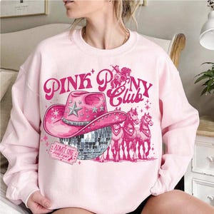 May include: A light pink sweatshirt with a graphic design featuring a pink cowboy hat, a disco ball, and the words "PINK PONY CLUB". The design also includes illustrations of horses and a ticket that says "ADMIT ONE".