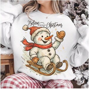 Op de afbeelding: Witte sweatshirt met een cartoon sneeuwpop met een rode sjaal en hoed, en bruine wanten. De sneeuwpop zit op een houten slee. De tekst "Believe in the magic of Christmas" staat erboven.