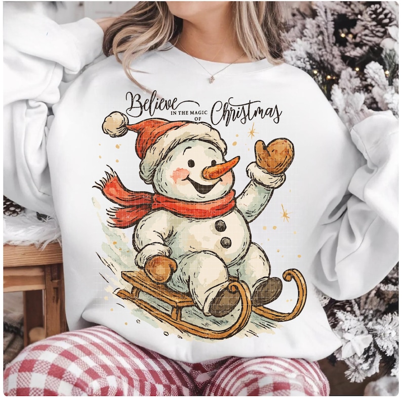 Op de afbeelding: Witte sweatshirt met een cartoon sneeuwpop met een rode sjaal en hoed, en bruine wanten. De sneeuwpop zit op een houten slee. De tekst "Believe in the magic of Christmas" staat erboven.
