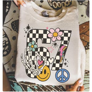 May include: A light beige sweatshirt with a colorful graphic design. The design includes a checkered number 67, a skeleton hand, flowers, a smiley face, a peace sign, and a lightning bolt. The sweatshirt is folded.