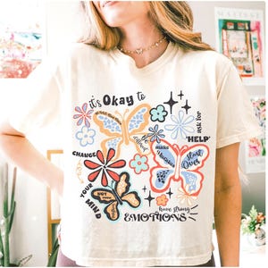 May include: A white t-shirt with a colorful graphic design featuring butterflies, flowers, and inspirational text. The text includes "it's Okay to", "CHANGE YOUR MIND", "ask for HELP", "Start Over", "Be different", "make Mistakes", and "have strong EMOTIONS".