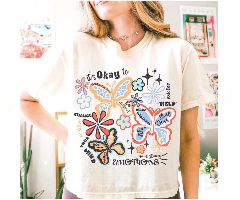 May include: A white t-shirt with a colorful graphic design featuring butterflies, flowers, and inspirational text. The text includes "it's Okay to", "CHANGE YOUR MIND", "ask for HELP", "Start Over", "Be different", "make Mistakes", and "have strong EMOTIONS".