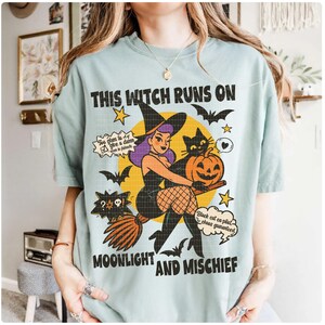 May include: A pale blue t-shirt with a Halloween graphic. The design shows a witch on a broom with a black cat and a pumpkin. The text on the shirt says "This Witch Runs On Moonlight And Mischief".