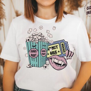 May include: White t-shirt with a graphic print of a bucket of popcorn with skulls, a ticket that says "Admit One Murder", and a vampire mouth. The popcorn bucket says "Fright Night" on the sides.