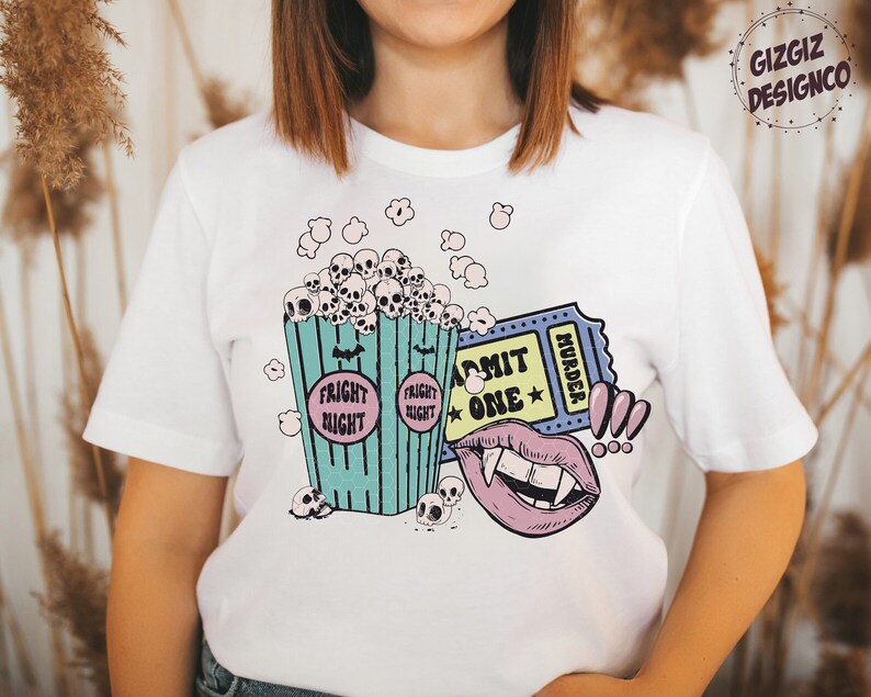 May include: White t-shirt with a graphic print of a bucket of popcorn with skulls, a ticket that says "Admit One Murder", and a vampire mouth. The popcorn bucket says "Fright Night" on the sides.