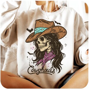 May include: Cream-colored sweatshirt featuring a graphic of a skull wearing a cowboy hat with the words "Long Live" and a purple bandana. The design includes bats and the word "Cowghouls".