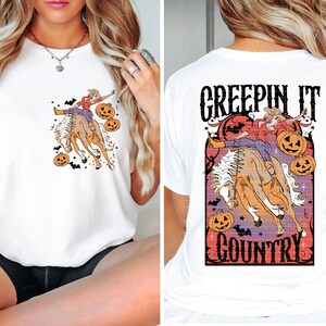 Puede incluir: Camiseta blanca con un gr&aacute;fico de una mujer montando un caballo con calabazas y murci&eacute;lagos. El texto "Creepin It Country" est&aacute; impreso en la parte posterior de la camiseta.